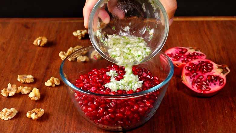A hand is seen pouring the dressing from a glass bowl into a large glass bowl half full of pomegranate arils. California walnuts kernels and an open pomegranate lie on the wooden counter in the background - Konkani Cucumber and Pomegranate Salad Recipe